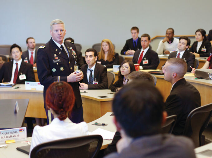 A lecturer dressed in decorated armed services uniform stands in front of a tiered desk auditorium talking to an audience