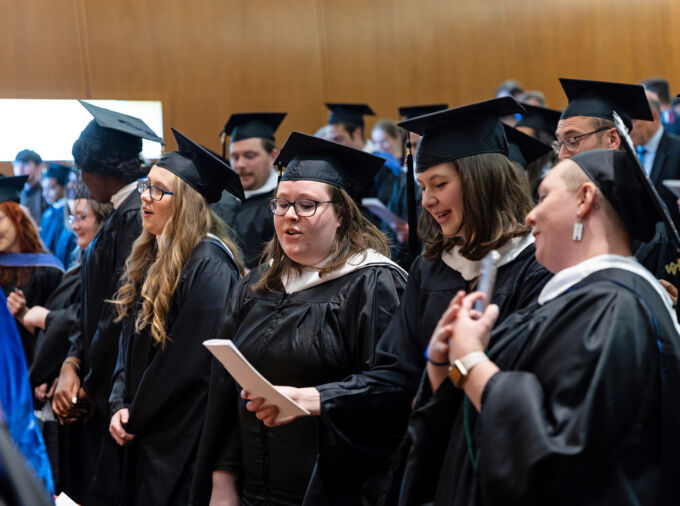 A roomful of graduates in dark cap and gown singing