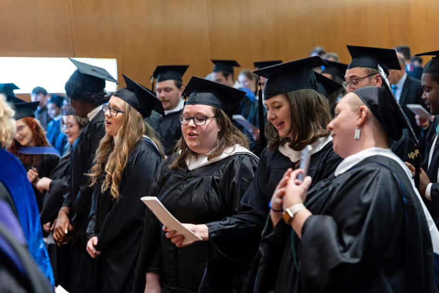 A roomful of graduates in dark cap and gown singing